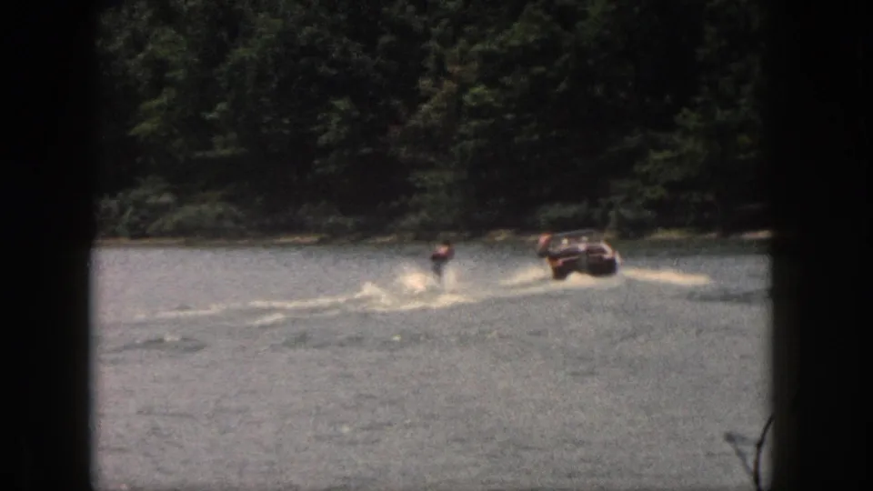 A distant skier and motorboat cross the wooded Michigan lake in a grainy 8mm home-movie frame.