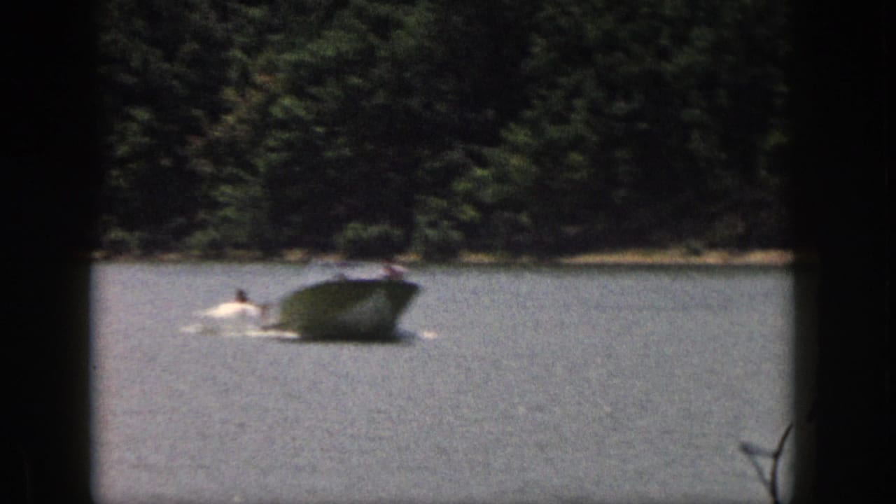 1961 Michigan 8mm frame showing a speedboat pulling a distant water skier across a lake bordered by dense trees.