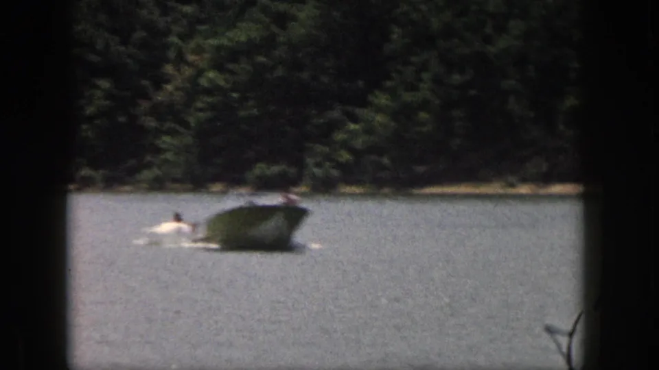 1961 Michigan 8mm frame showing a speedboat pulling a distant water skier across a lake bordered by dense trees.