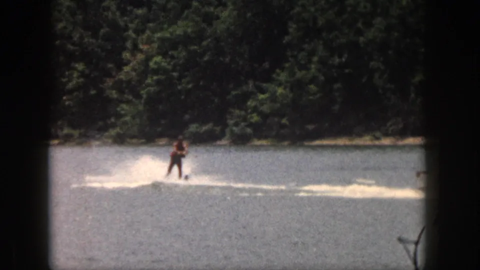 1961 Michigan 8mm frame showing a man balancing on water skis with spray behind him on a wooded lake.