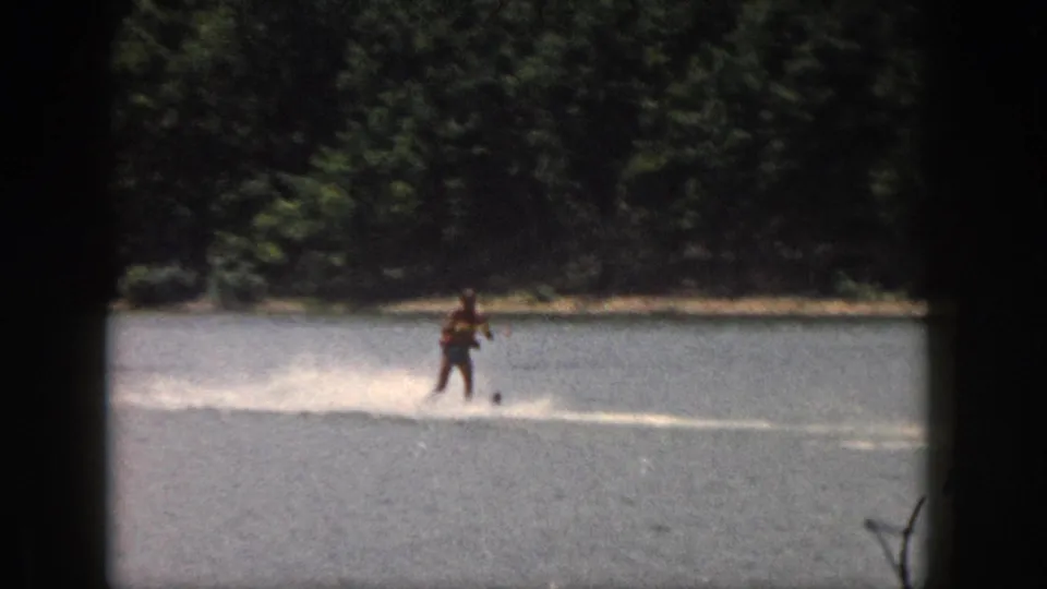 1961 Michigan home-movie frame of a water skier moving across open lake water with a wooded shoreline in back.