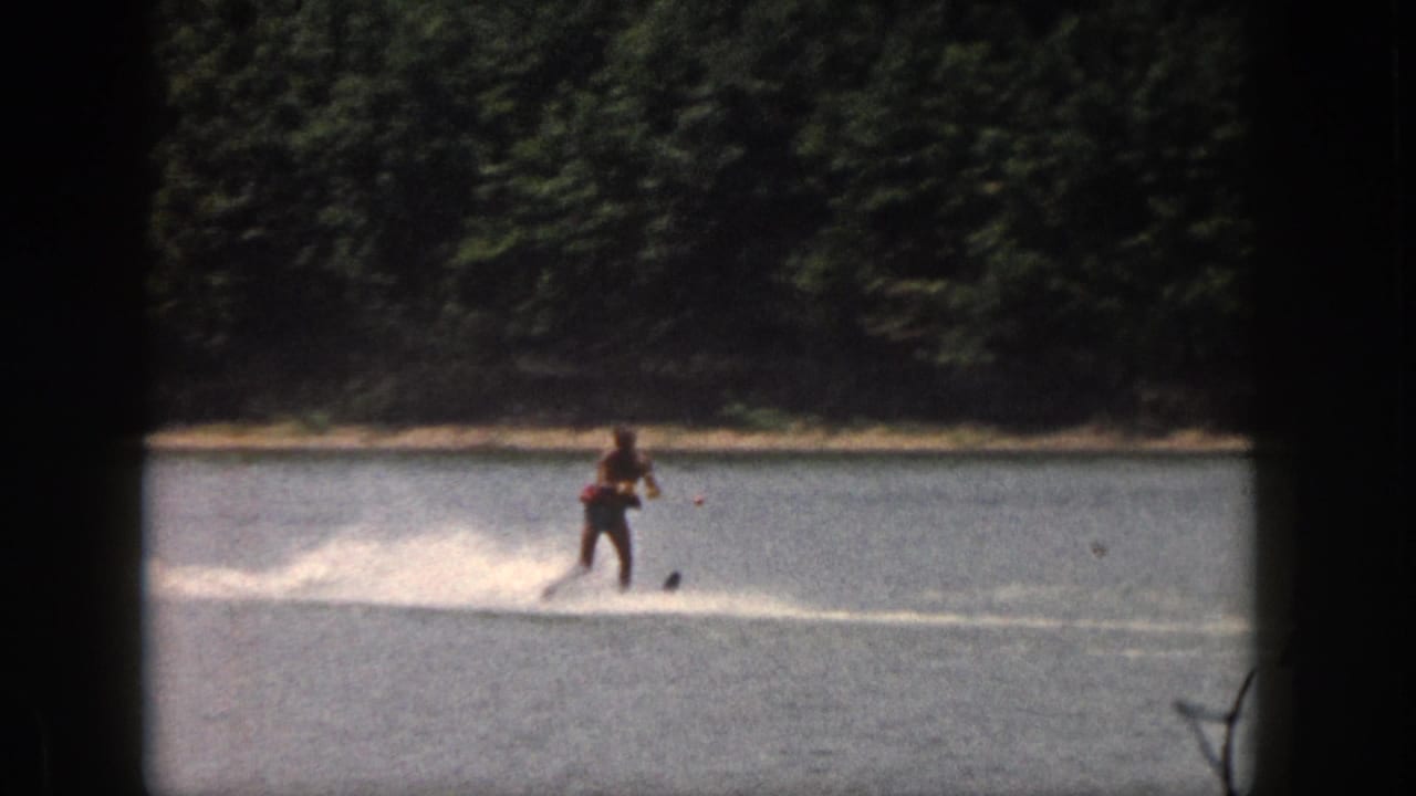 1961 Michigan 8mm frame showing a distant water skier leaving a white spray trail on a calm lake near trees.