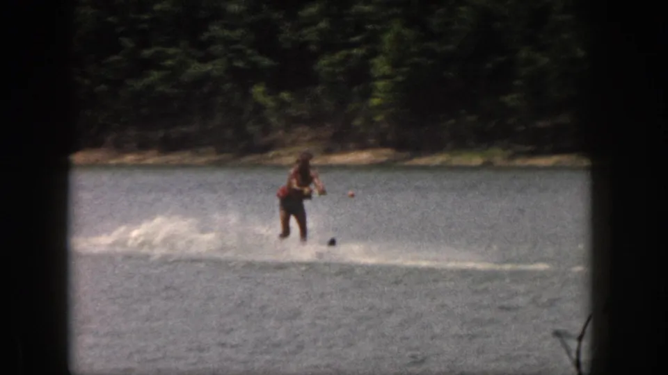 A skier in a life vest glides across a Michigan lake in 1961, framed by dark 8mm film borders and shoreline trees.
