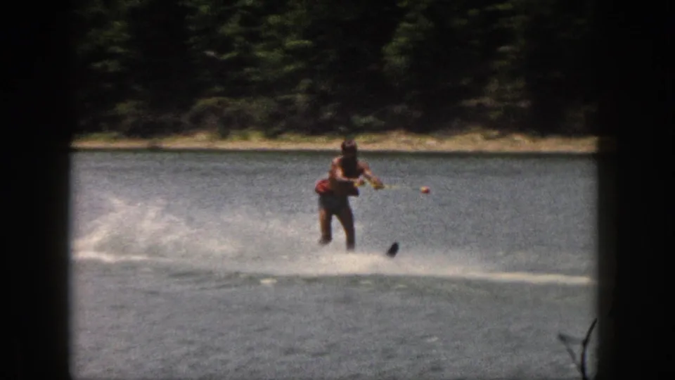 Closer 8mm view of the water skier holding the tow rope while spray rises from a single ski on a wooded Michigan lake.