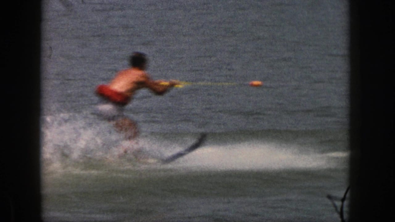 1961 Michigan home-movie frame showing a shirtless man in red trunks water skiing on a lake with a tow rope and wake spray.