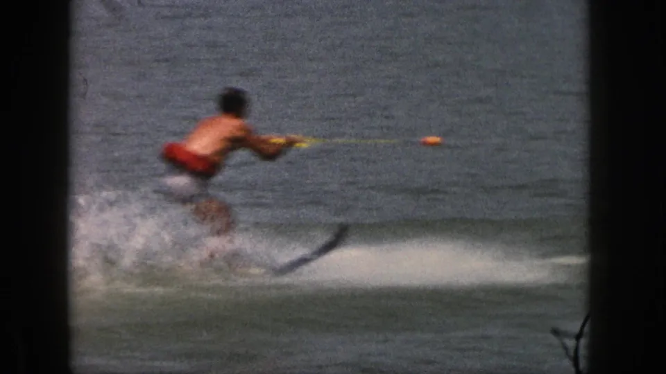 1961 Michigan home-movie frame showing a shirtless man in red trunks water skiing on a lake with a tow rope and wake spray.