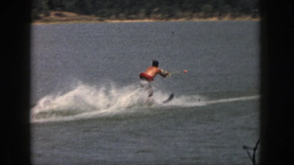A shirtless man in red trunks skis across a Michigan lake, with white spray, a tow rope, and a distant wooded shoreline visible.
