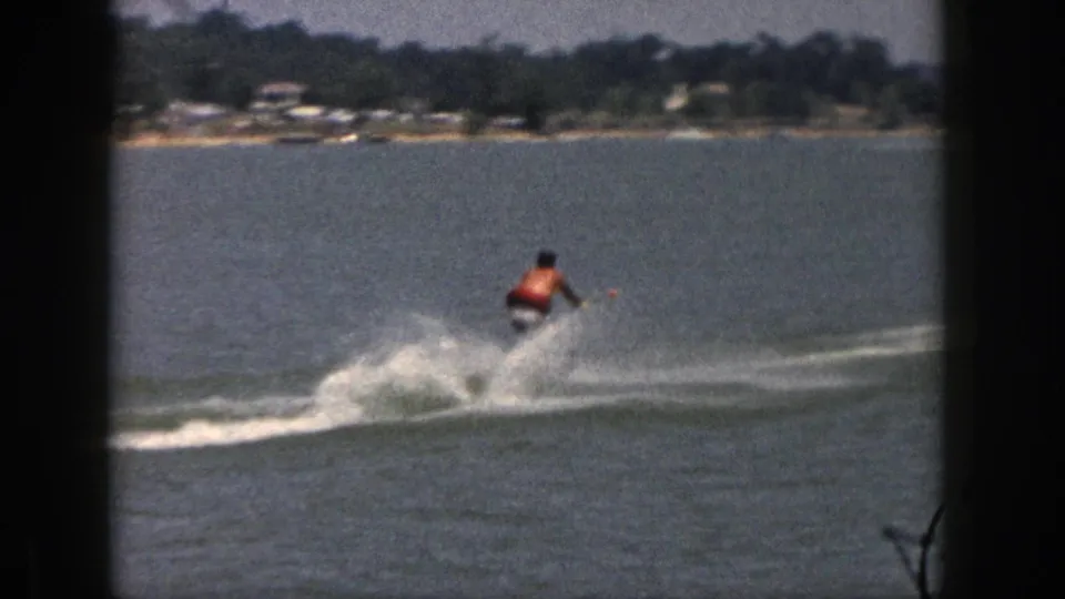 The skier moves farther away across a Michigan lake, leaving a wake with trees and lakeside houses along the distant shoreline.