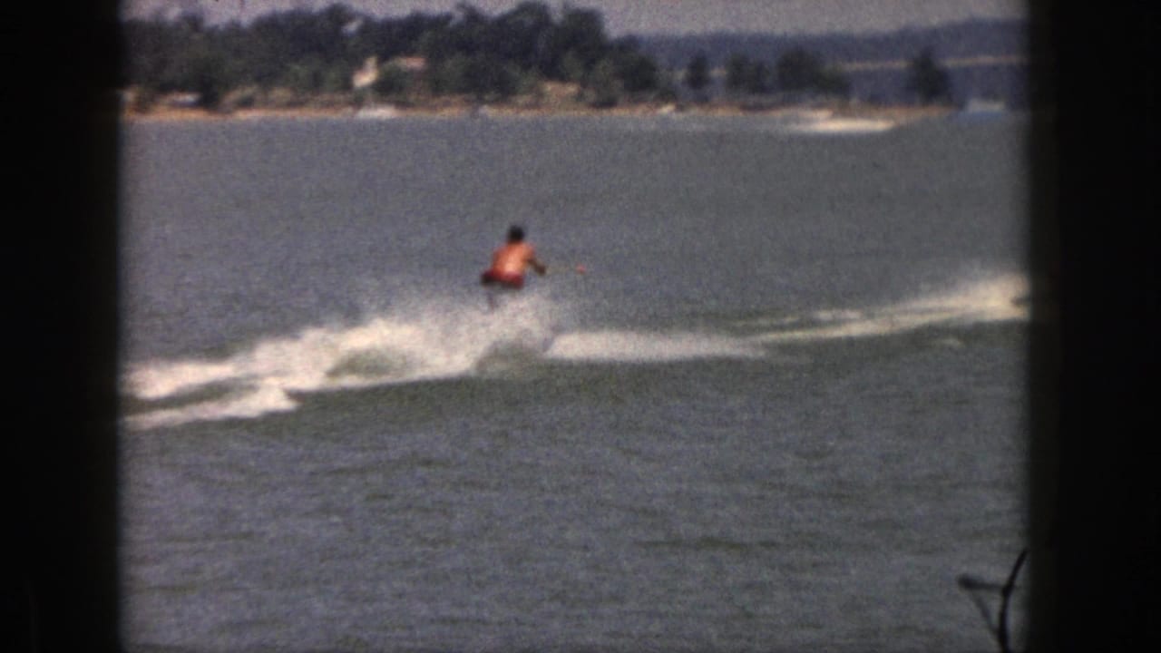 1961 Michigan 8mm frame of a man water-skiing across a lake, seen through dark window framing with a distant shoreline and bridge.