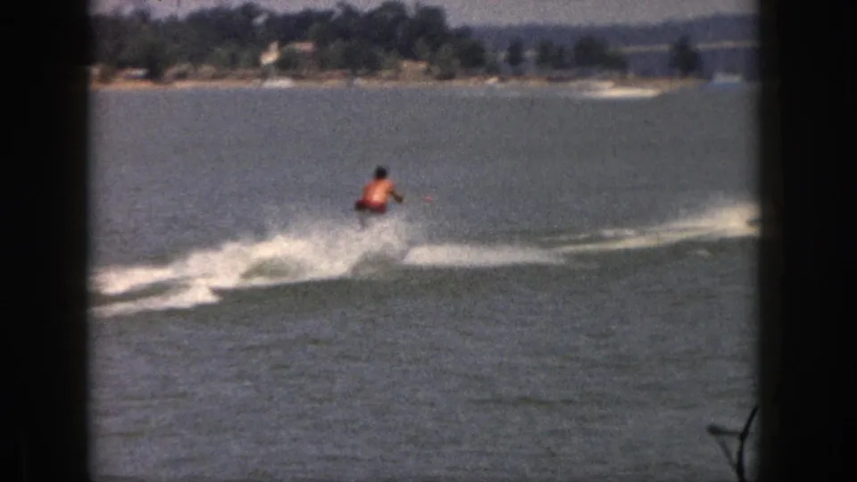 1961 Michigan 8mm frame of a man water-skiing across a lake, seen through dark window framing with a distant shoreline and bridge.
