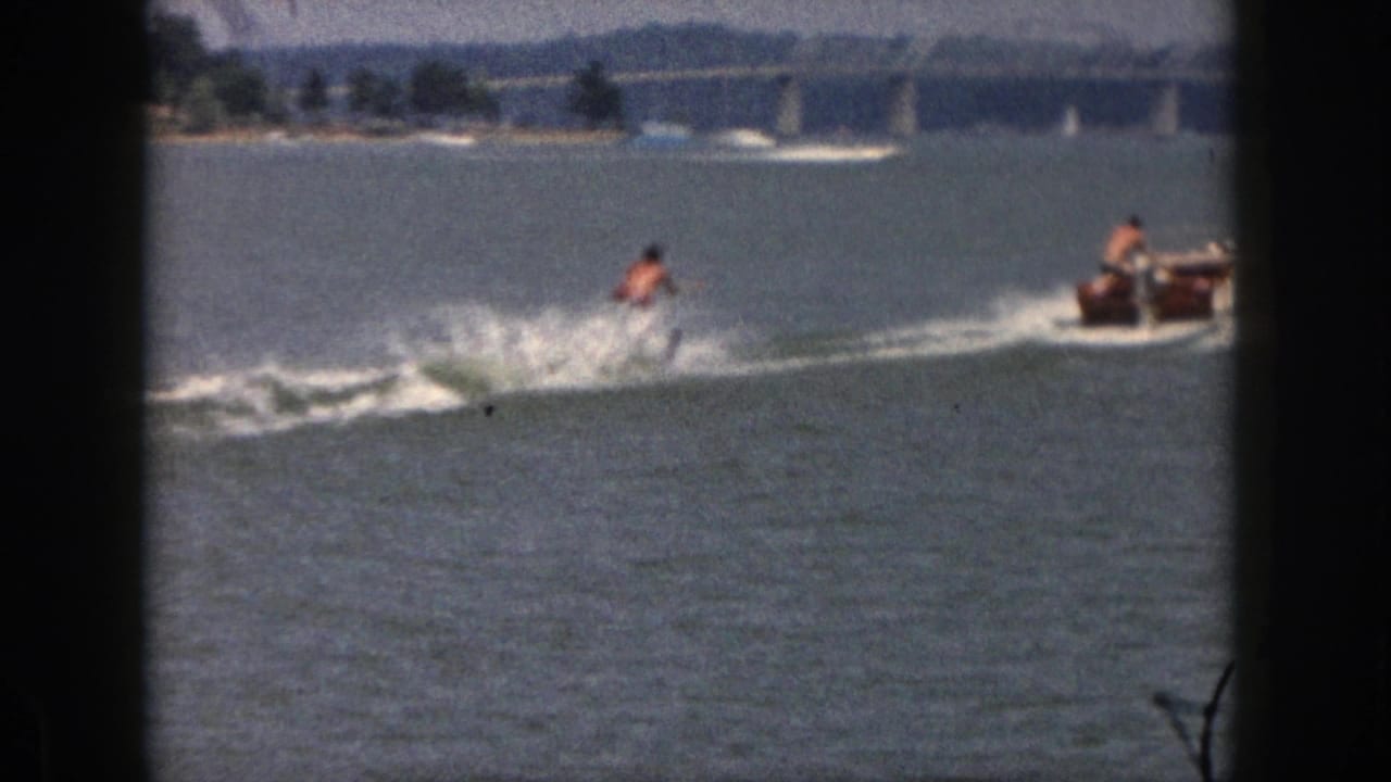 The skier continues across the Michigan lake as spray rises and a small motorboat appears ahead near the distant bridge.