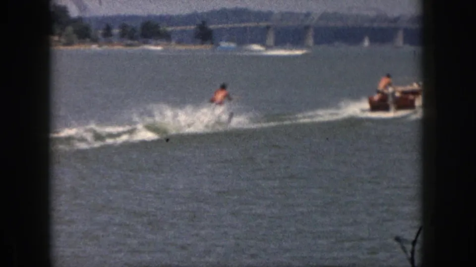 The skier continues across the Michigan lake as spray rises and a small motorboat appears ahead near the distant bridge.