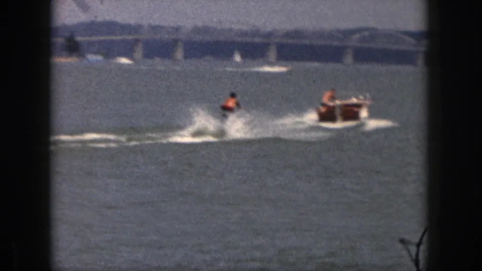 A man water-skiing in 1961 Michigan splashes low on the lake near the tow boat, with bridge spans across the background.