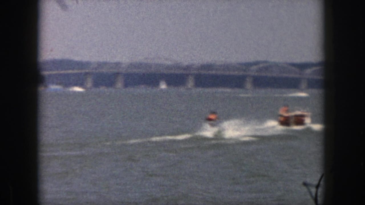 1961 Michigan 8mm frame showing a distant water-skier pulled by a small motorboat across open water near a long bridge.