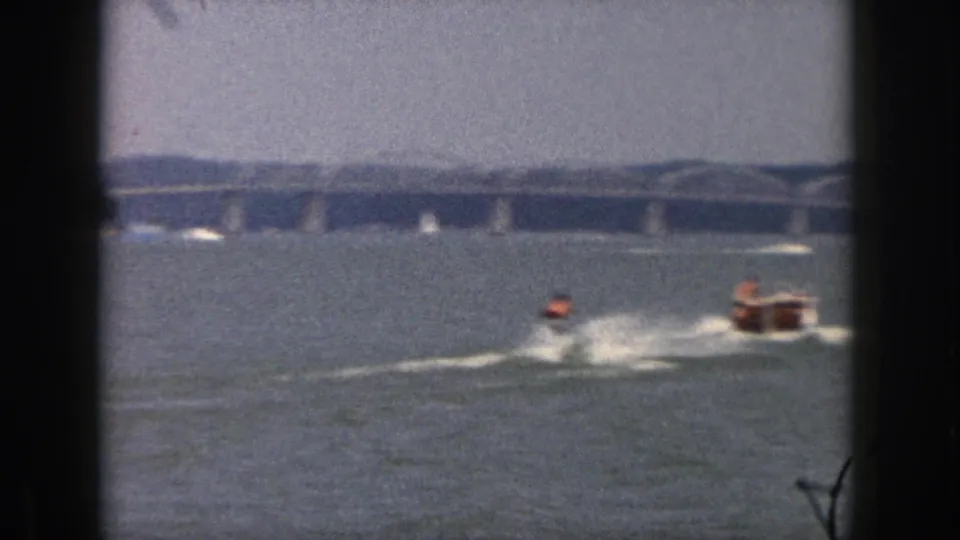 1961 Michigan 8mm frame showing a distant water-skier pulled by a small motorboat across open water near a long bridge.