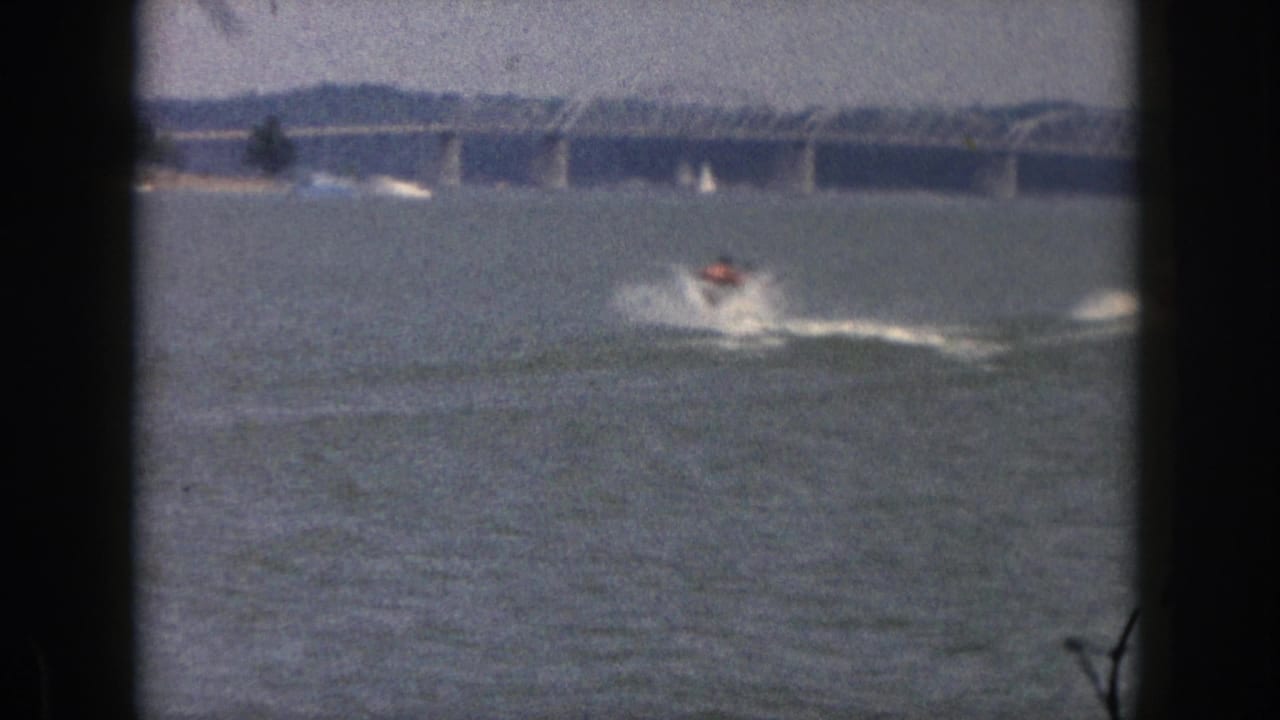 A distant water-skier moves across the water, leaving spray below a long bridge.