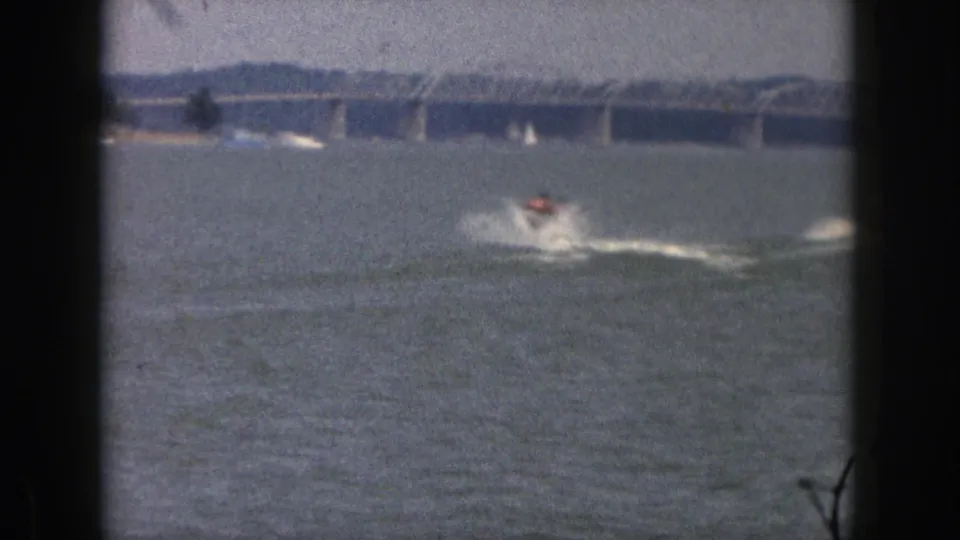 1961 Michigan 8mm frame with a distant water-skier in an orange vest moving across open water below a long bridge.