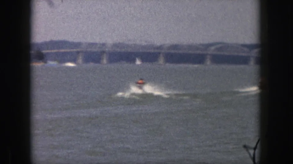1961 Michigan home-movie frame showing a water-skier crossing choppy water, with a long bridge and sailboats in the distance.