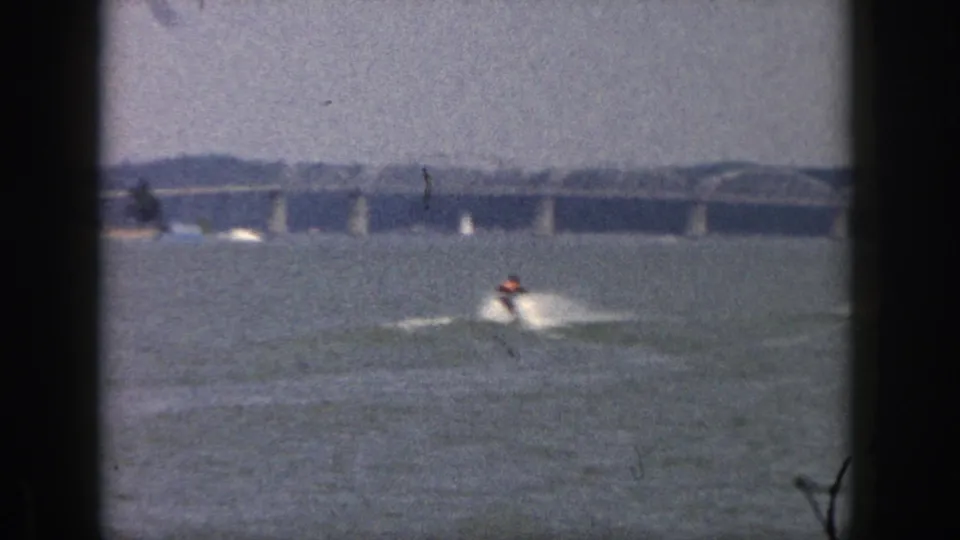 1961 Michigan 8mm frame showing a distant water-skier throwing spray across open water with bridge spans behind.