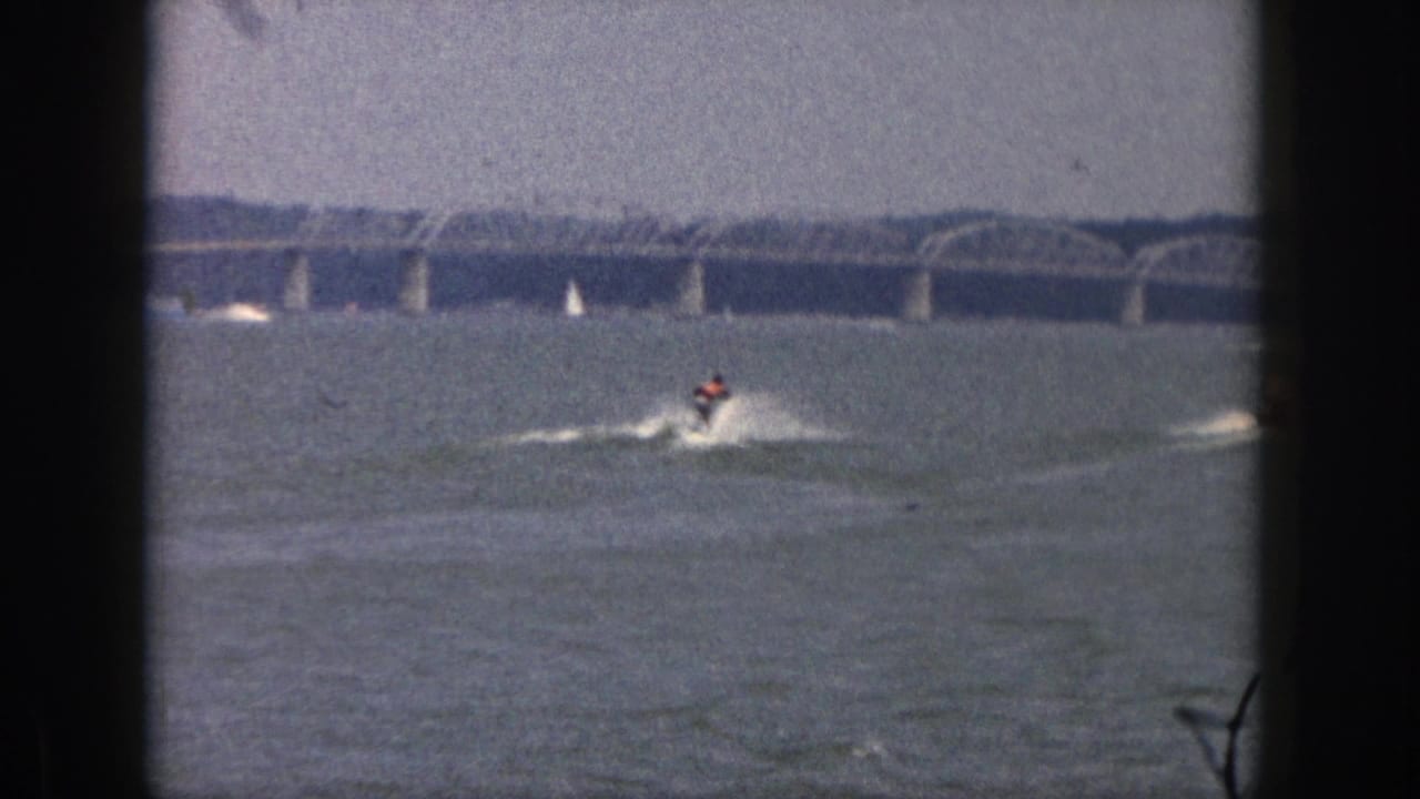 1961 Michigan 8mm frame showing a distant water skier being towed across a lake beneath a hazy sky and long arched bridge.