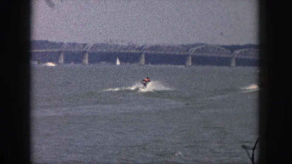 1961 Michigan 8mm frame showing a distant water skier being towed across a lake beneath a hazy sky and long arched bridge.