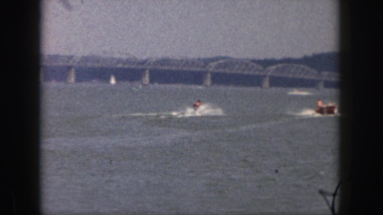 Water skier and tow wake appear on a lake near a small boat and distant bridge.