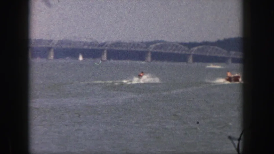 1961 Michigan home-movie frame with a skier towed across the lake as a small boat appears near the right edge and a bridge spans the distance.