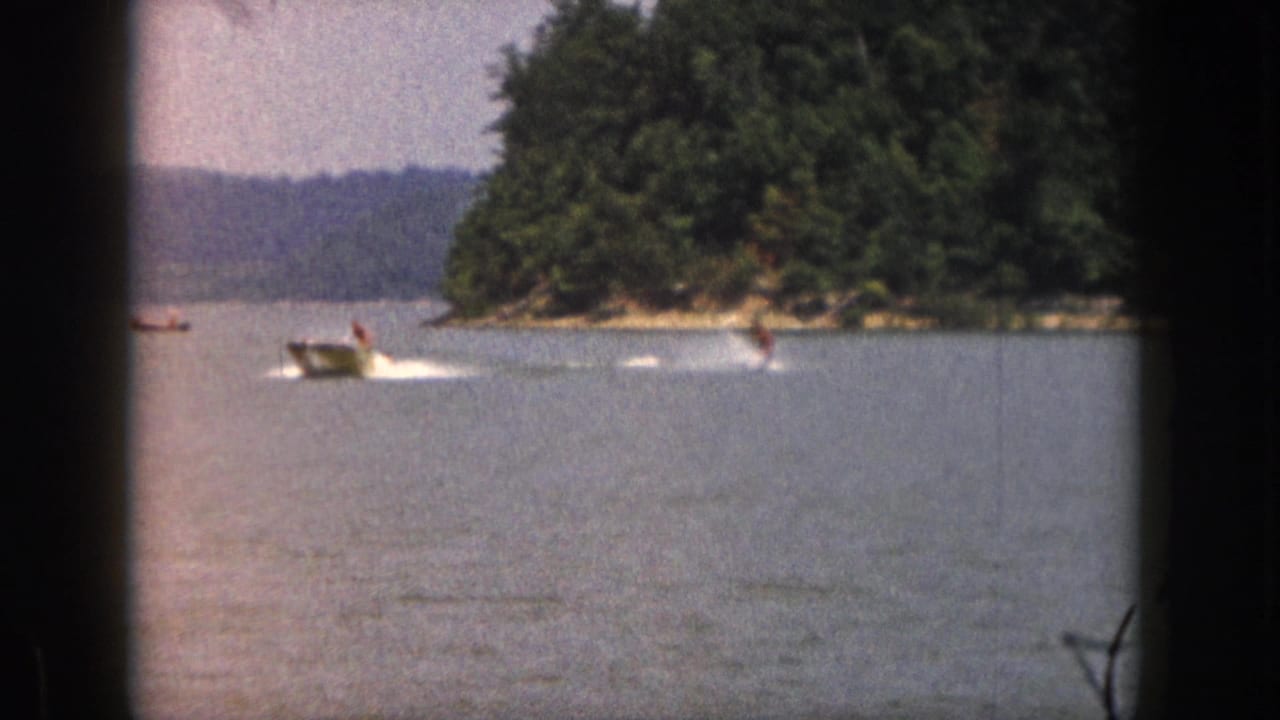 1961 Michigan home-movie frame showing a motorboat pulling a distant water skier across a lake near trees.