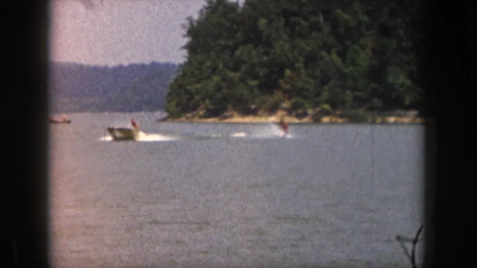 1961 Michigan home-movie frame showing a motorboat pulling a distant water skier across a lake near trees.