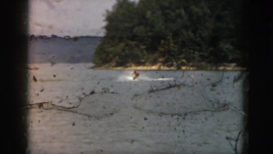 Home-movie frame from 1961 Michigan showing a distant skier cutting across lake water near a wooded bank.