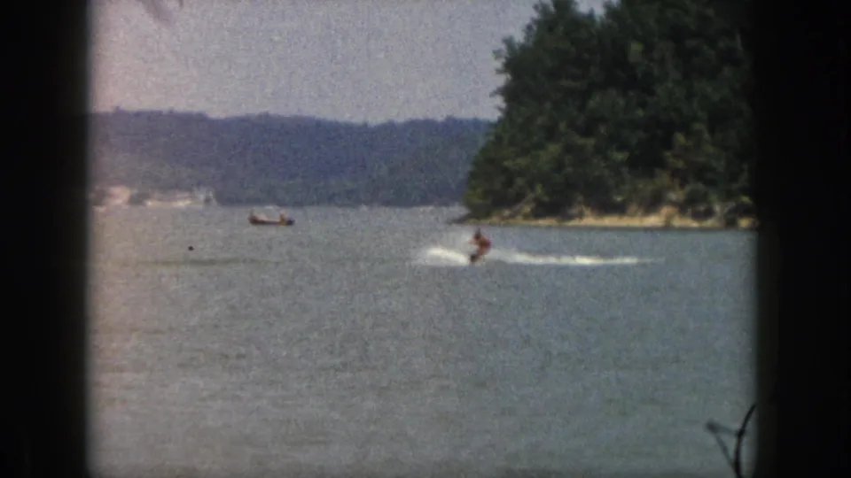 1961 Michigan vacation footage frame with a water skier making a white wake near a forested shoreline.