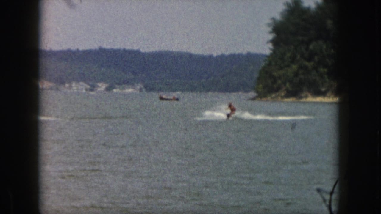 1961 Michigan 8mm frame of a distant water-skier being pulled across a lake near a wooded shore.