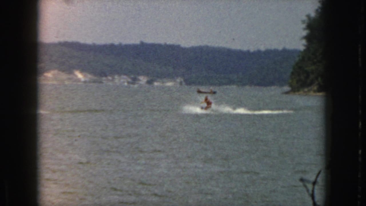 Distant water-skier and boat on a lake, framed by dark 8mm borders and shoreline trees.