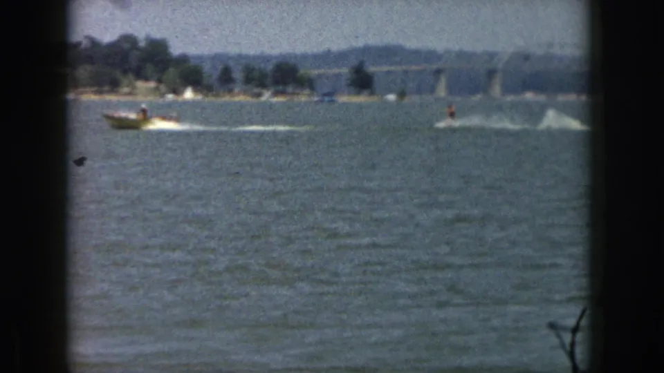 1961 Michigan 8mm frame with a motorboat and distant water-skier moving across a lake near a wooded bank.