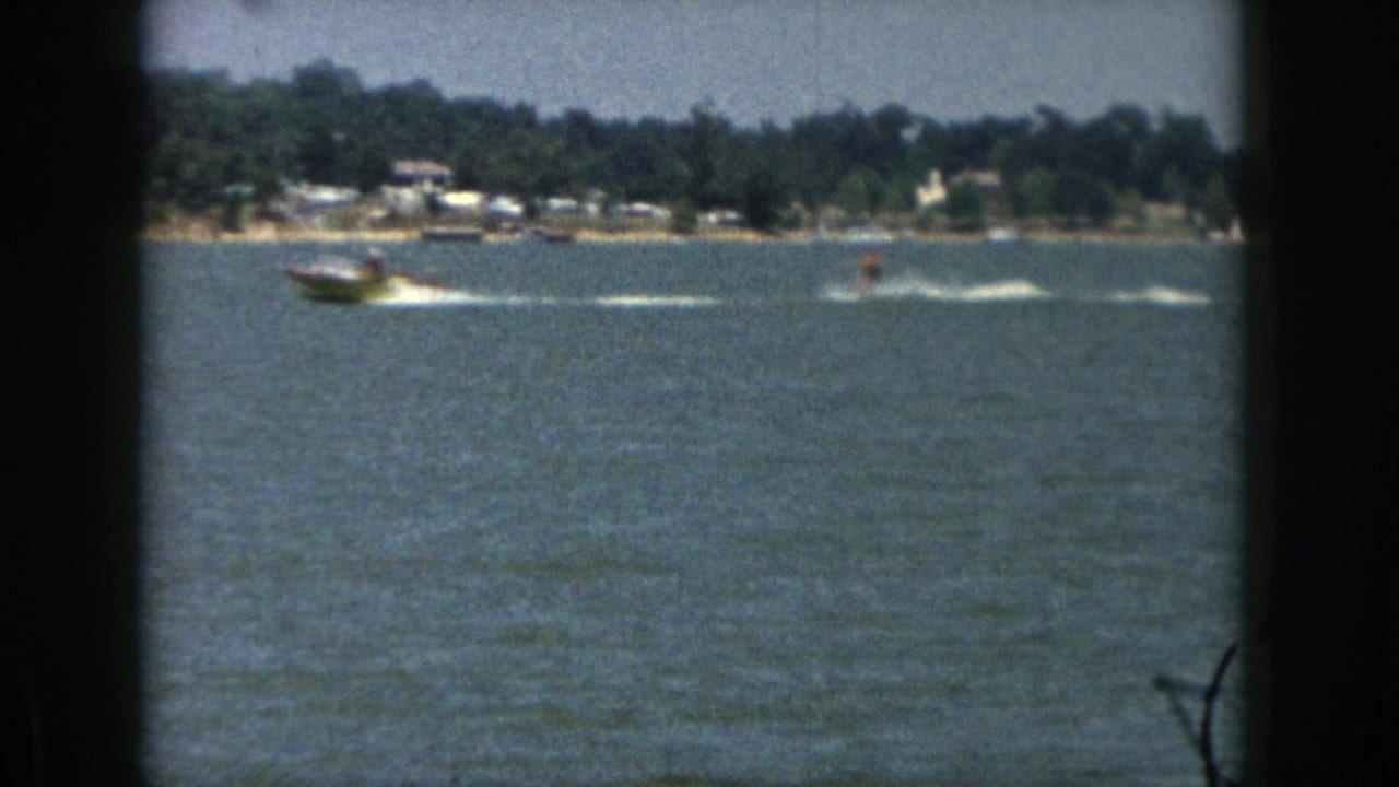 1961 Michigan 8mm frame of a motorboat pulling a distant water skier across a lake with trees and small houses on shore.