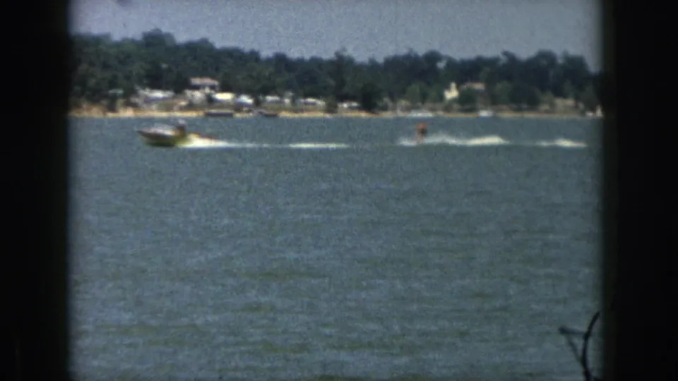 1961 Michigan 8mm frame of a motorboat pulling a distant water skier across a lake with trees and small houses on shore.