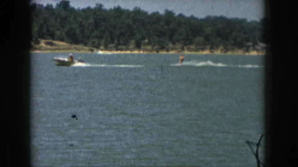 Distant 1961 Michigan lake scene with a motorboat towing a water skier past a wooded shore in soft 8mm color.