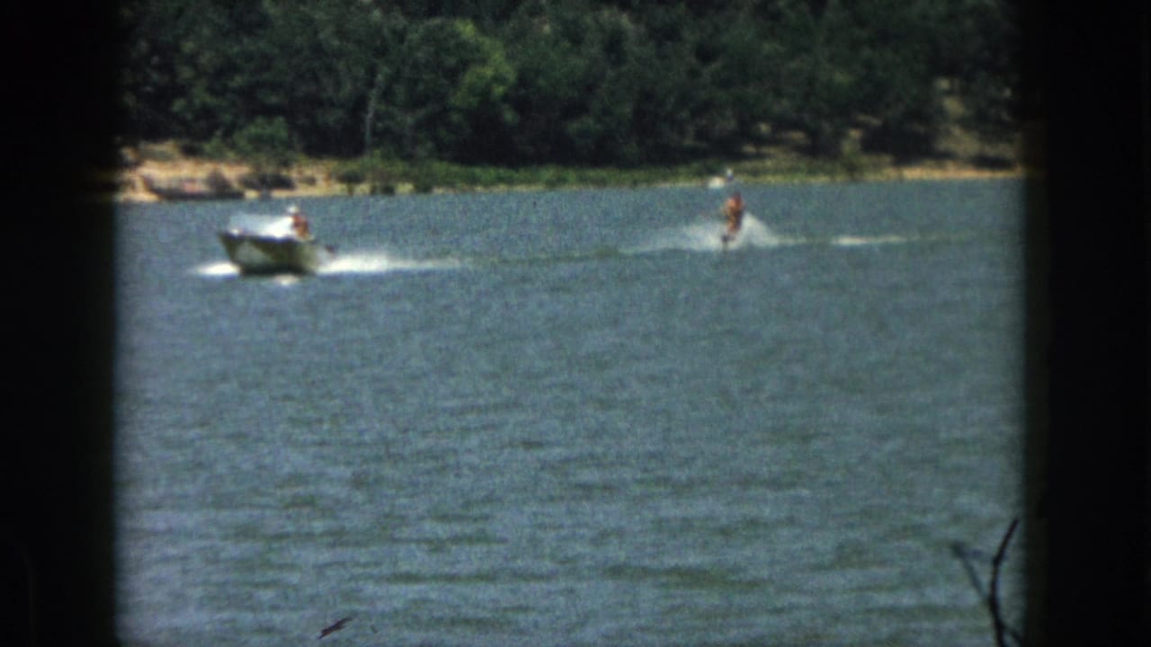1961 Michigan home-movie frame showing a motorboat towing a distant man on water skis across a tree-lined lake.