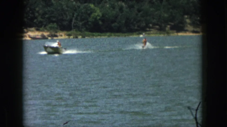 1961 Michigan home-movie frame showing a motorboat towing a distant man on water skis across a tree-lined lake.