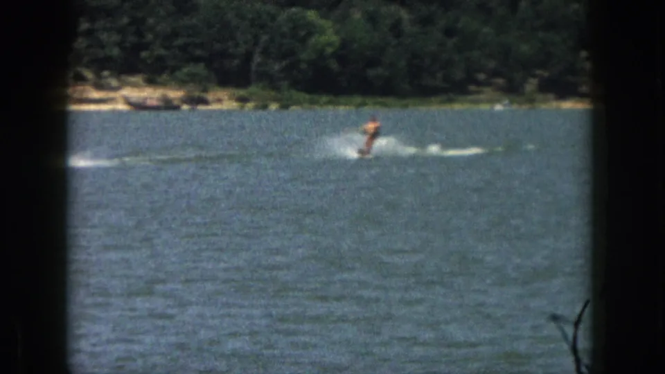 Distant 1961 Michigan home-movie view of a man water skiing across a lake, with spray and a wooded shore in the background.