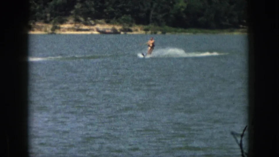 1961 Michigan frame showing a distant male water skier gliding across open water with a long wake and wooded shoreline beyond.