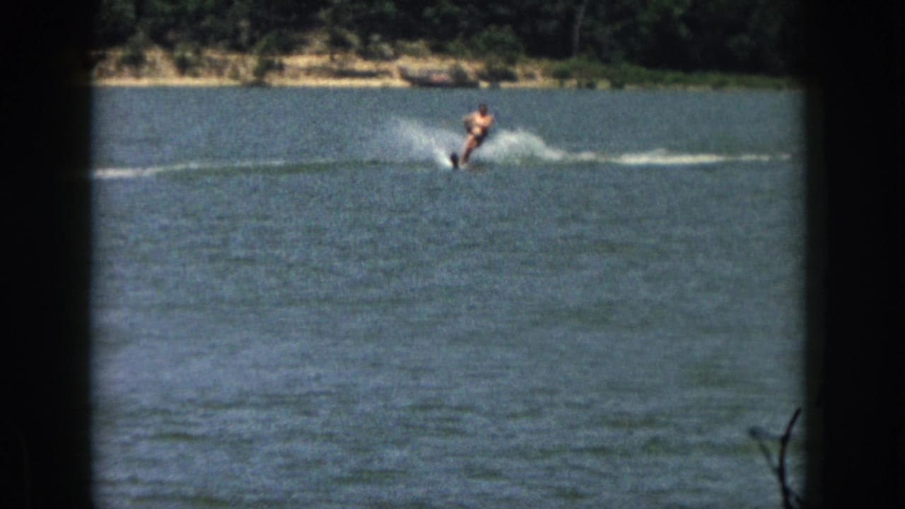 1961 Michigan 8mm frame of an adult man water-skiing across blue lake water near a tree-lined sandy shore.