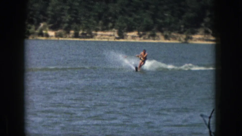1961 Michigan 8mm still of an adult man leaning on water skis over blue lake water near a wooded bank.