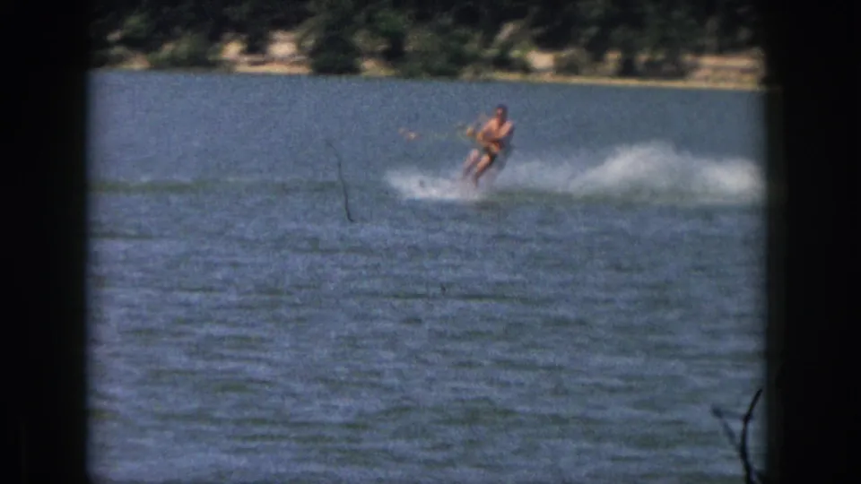 1961 Michigan home-movie frame of a distant adult water skier crossing the lake with a broad white spray plume.