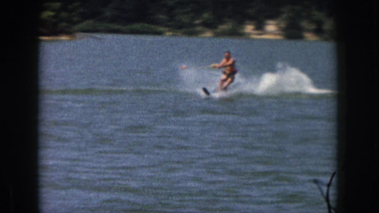 1961 Michigan home-movie frame showing a man water skiing on a lake, with white spray trailing behind him and a wooded shoreline in the distance.