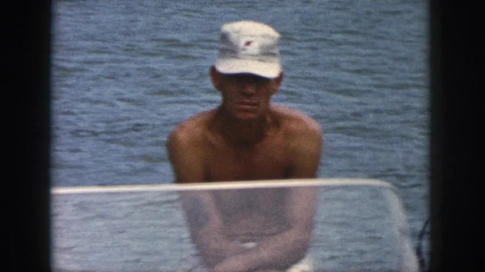 1961 Michigan home-movie frame showing a shirtless man in a white cap seated behind a low boat windshield on the lake.