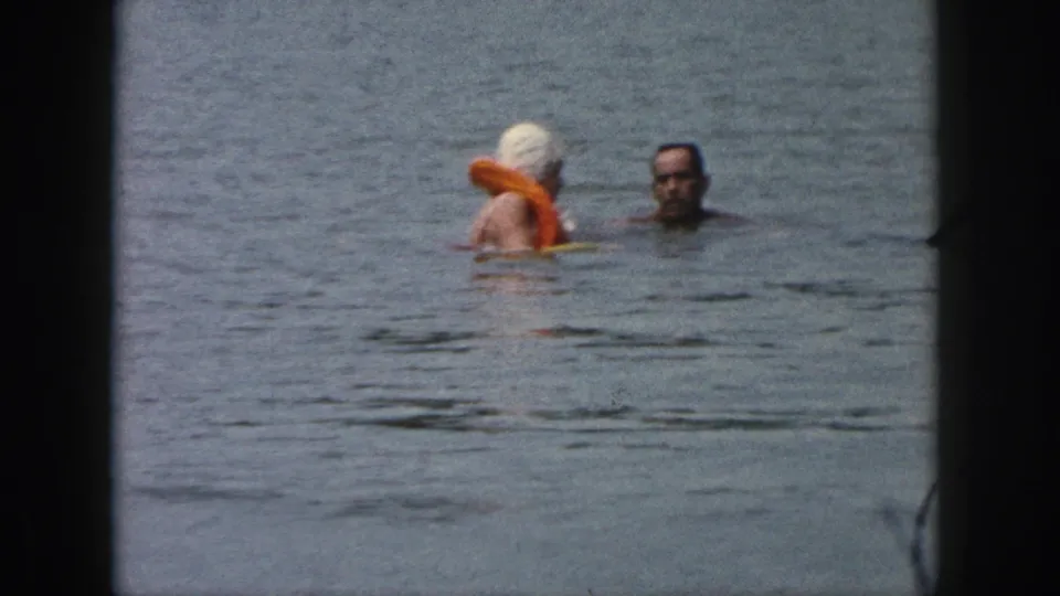 1961 Michigan lake frame showing two people in the water, including one in an orange life vest and another swimmer facing the camera.