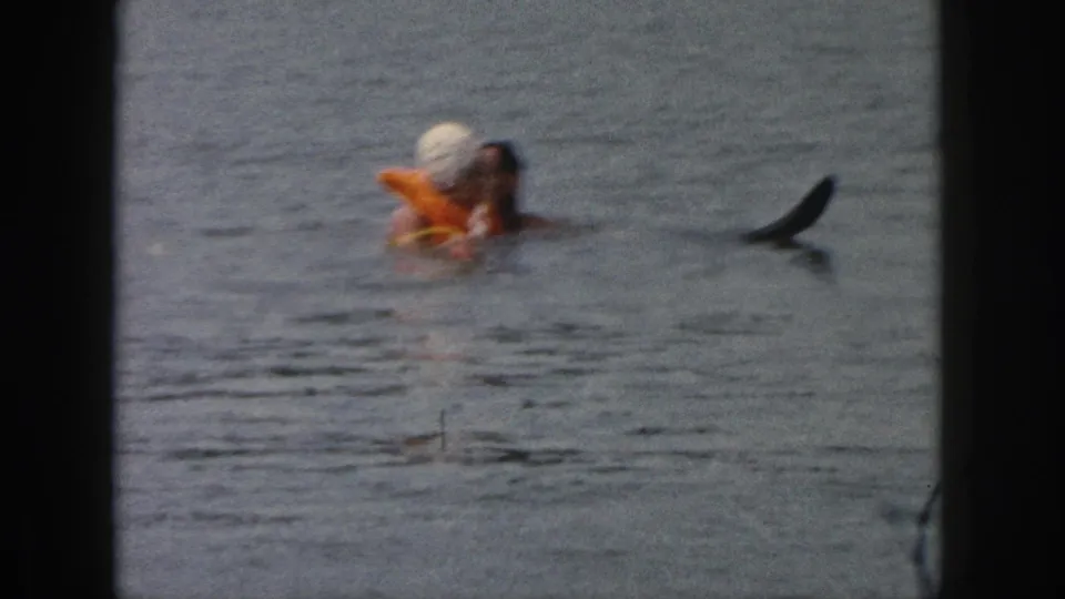 1961 Michigan home-movie frame of two adults close together in a lake, with the orange float prominent and a dark shape at right.