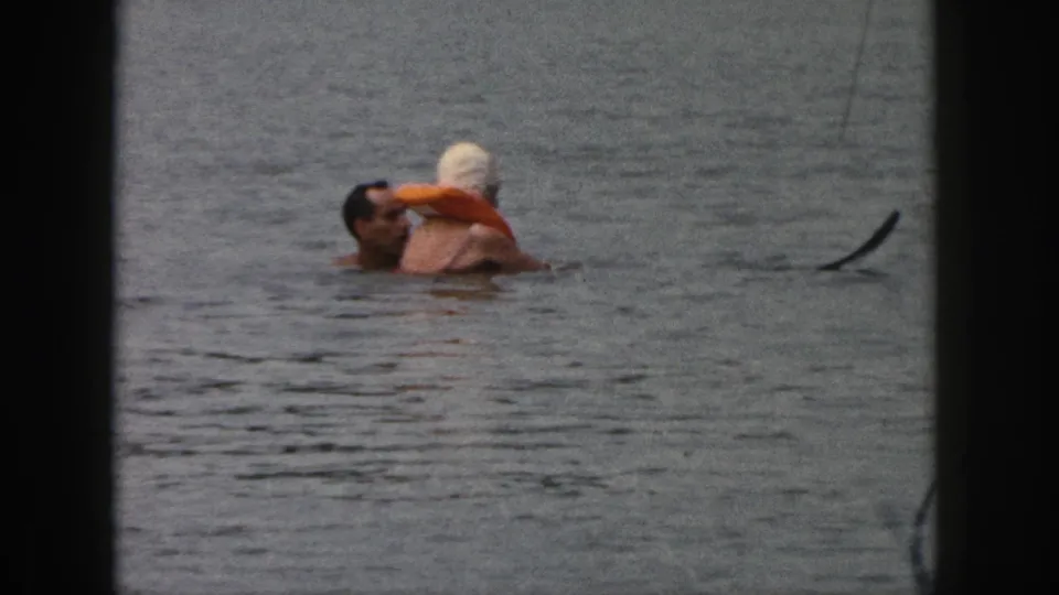 1961 Michigan home-movie frame of a man holding or supporting a woman wearing a swim cap and orange float in lake water.