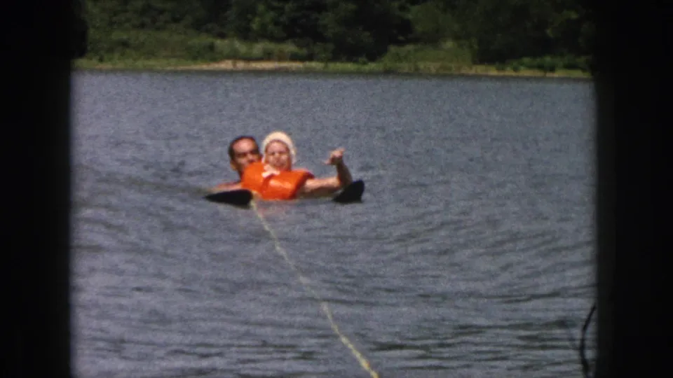 1961 Michigan home-movie frame showing the pair floating on a lake, one arm raised near the visible tow rope and ski.